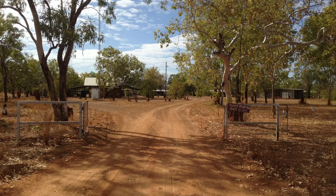 A dirt road leads through a gate. Near the gate are trees and beyond that are more trees and buildings.