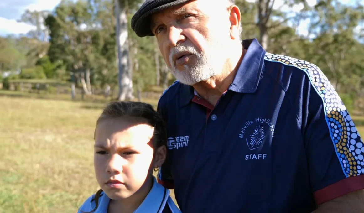 Elderly Aboriginal man in blue polo shirt with Aboriginal designs on the sleeves and wearing a cap stands with a young girls in a blue shirt. In the background is grass and trees.