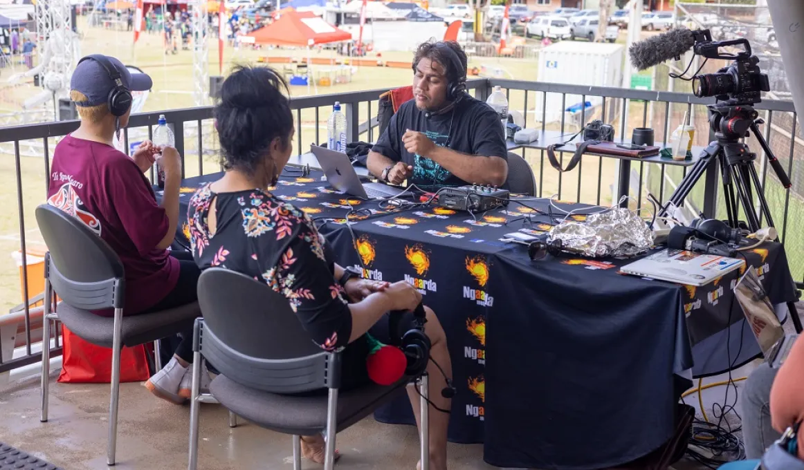 Three people sit around a table covered with a large mainly black cloth. Next to the table is a camera and microphone on a tripod. On the table are laptops and other paraphernalia. In the background are people, cars and buildings.