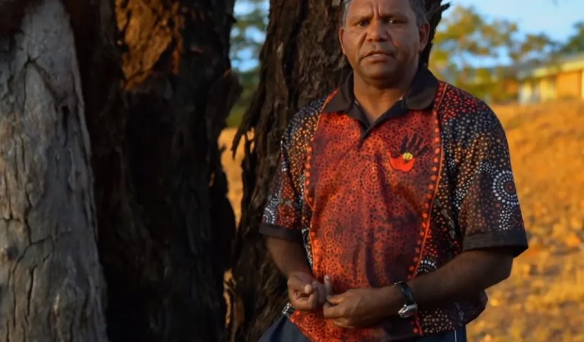 An Aboriginal man with short hair and wearing a black and orange polo necked shirt looks at camera.