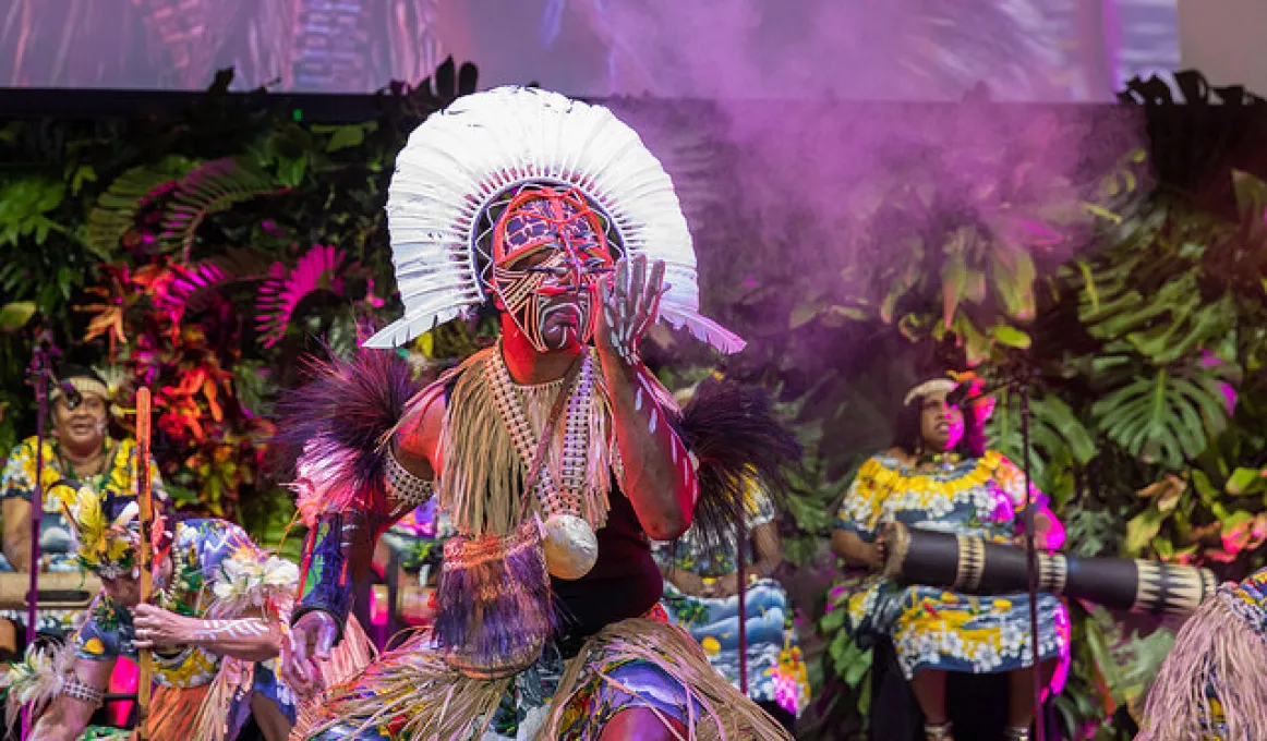 Dancer dressed in traditional Torres Strait Islander ceremonial clothing in the foreground with musicians in similar wear in background.