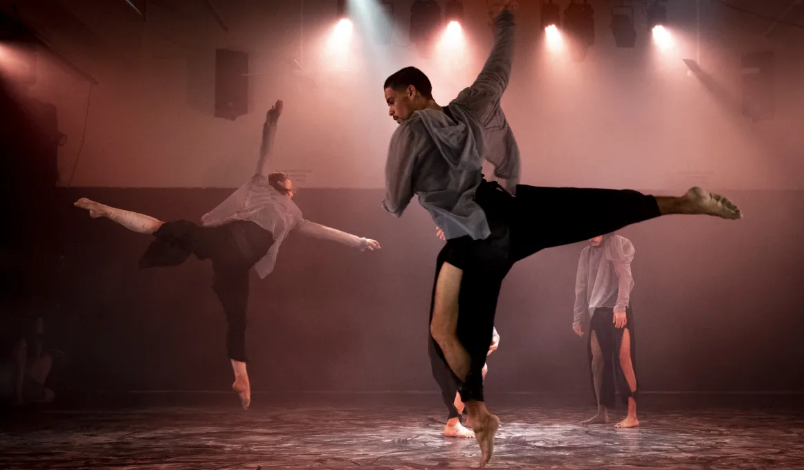 Four male and female dancers on a floor in softly lit room. Two dancers are in action while two others stand and crouch.