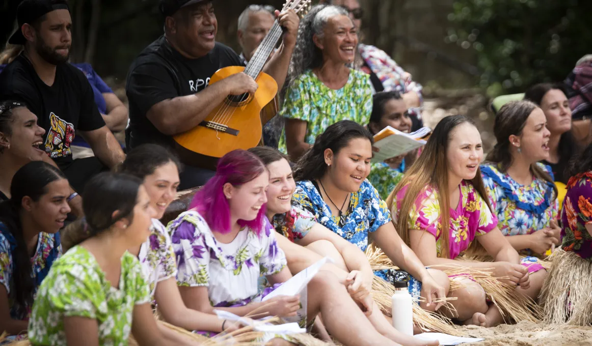 A group of people sit on chairs or on the ground and most wear colourful clothing. One man holds a guitar while some others hold papers. Most have their mouths open. In the background are trees.