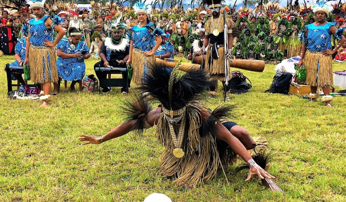 A dancer in traditional Torres Strait Islander dance wear squats on grass looking at a white round shaped object. In the background are many people dressed in dance costumes, trees and sky.