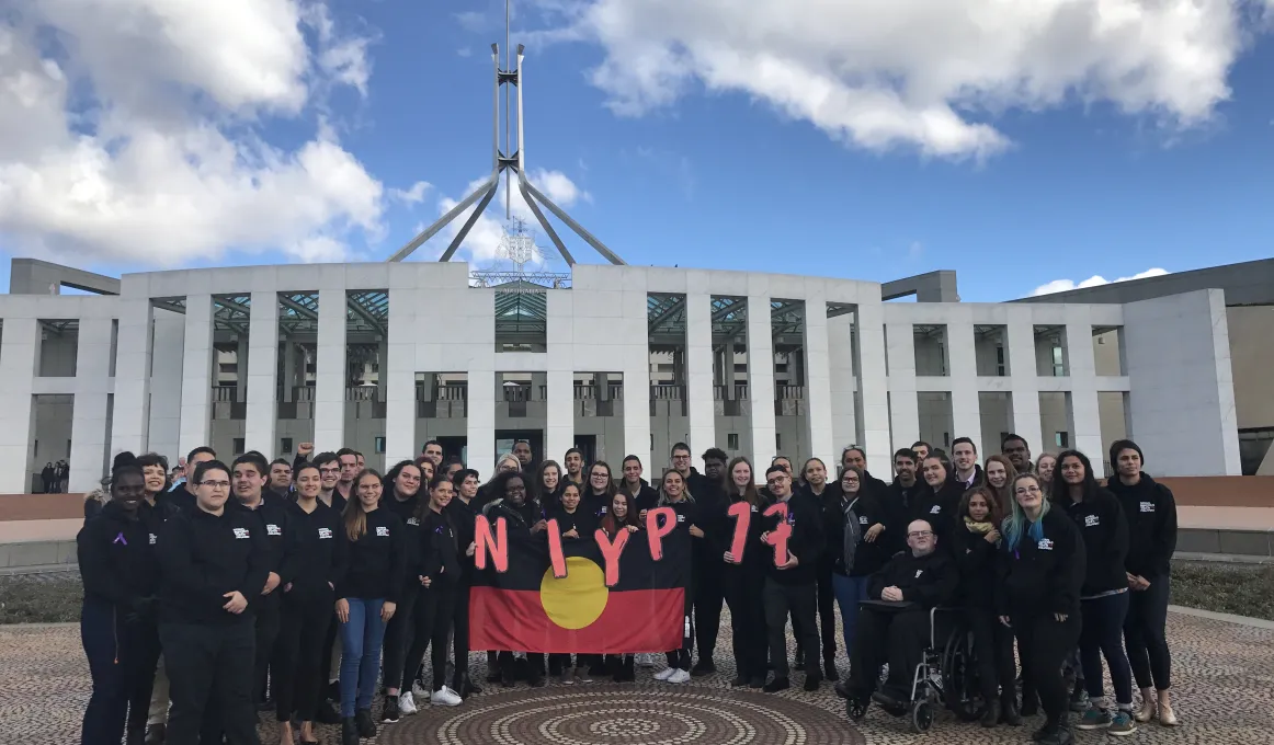 Group of Indigenous young adults dressed mainly in black tops stand in front of the Australian Parliament House holding the Aboriginal flag and the letters and numbers N.I.Y.P. 17