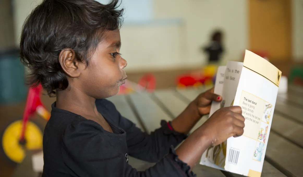 Pictured is an Indigenous school aged boy in a classroom reading a school book.