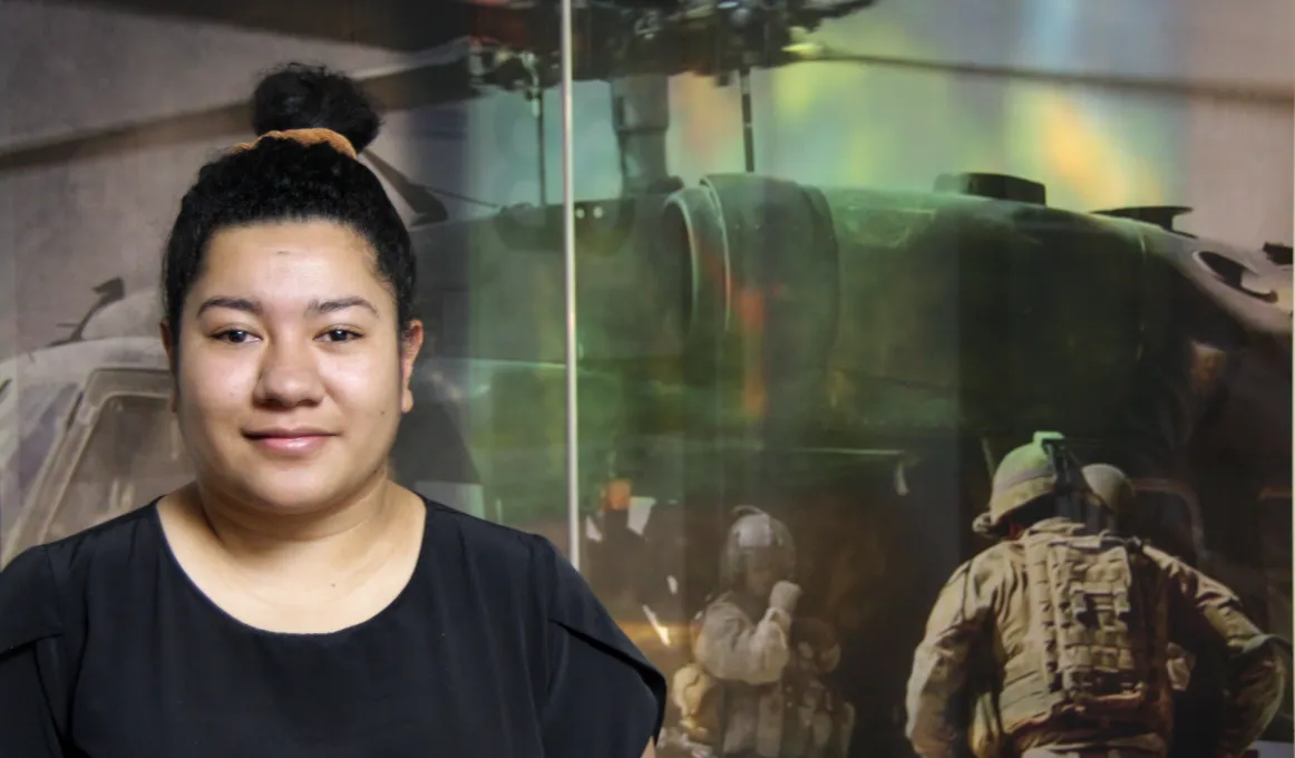 A woman wearing a black shirt with her hair in a knot. In the background is the image of a helicopter with military personnel in front of it.