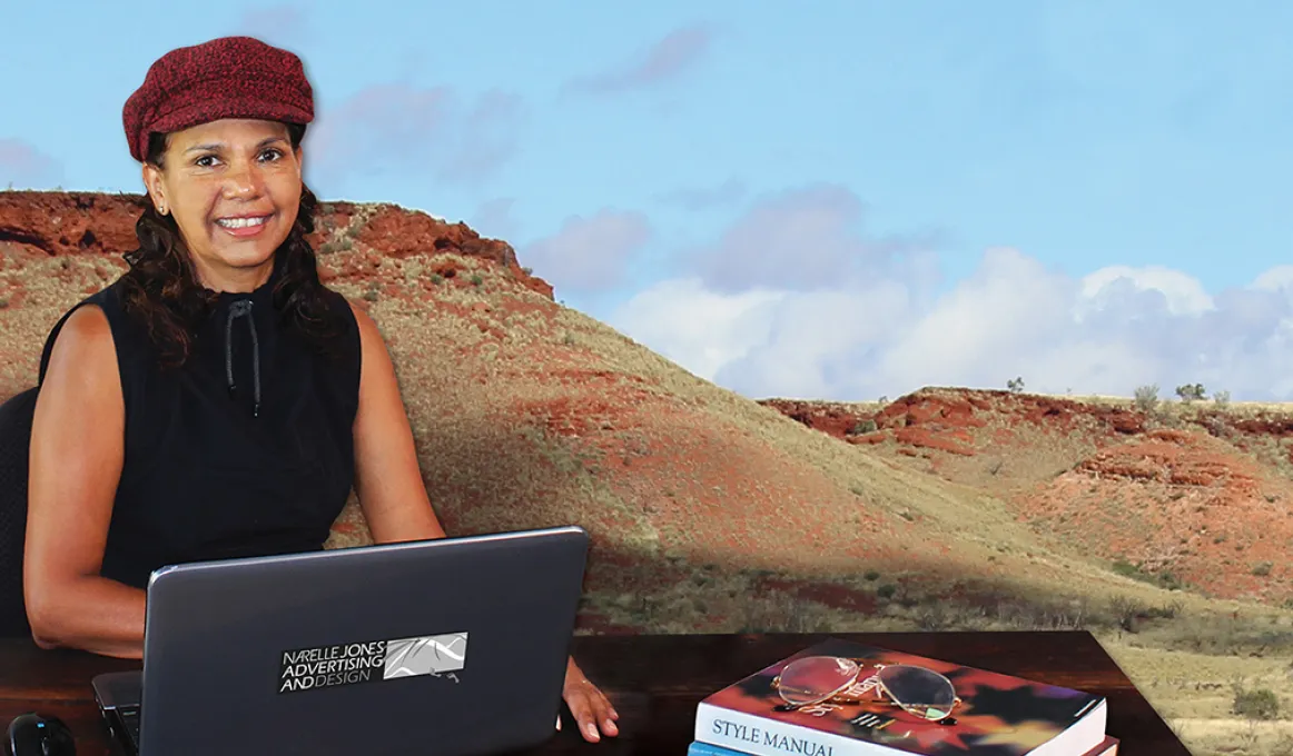 A woman in black top and red cap sits at a desk on which is a laptop and two books. In the background is a large photo of a dry landscape comprising two hills ochre red and pale green in colour.