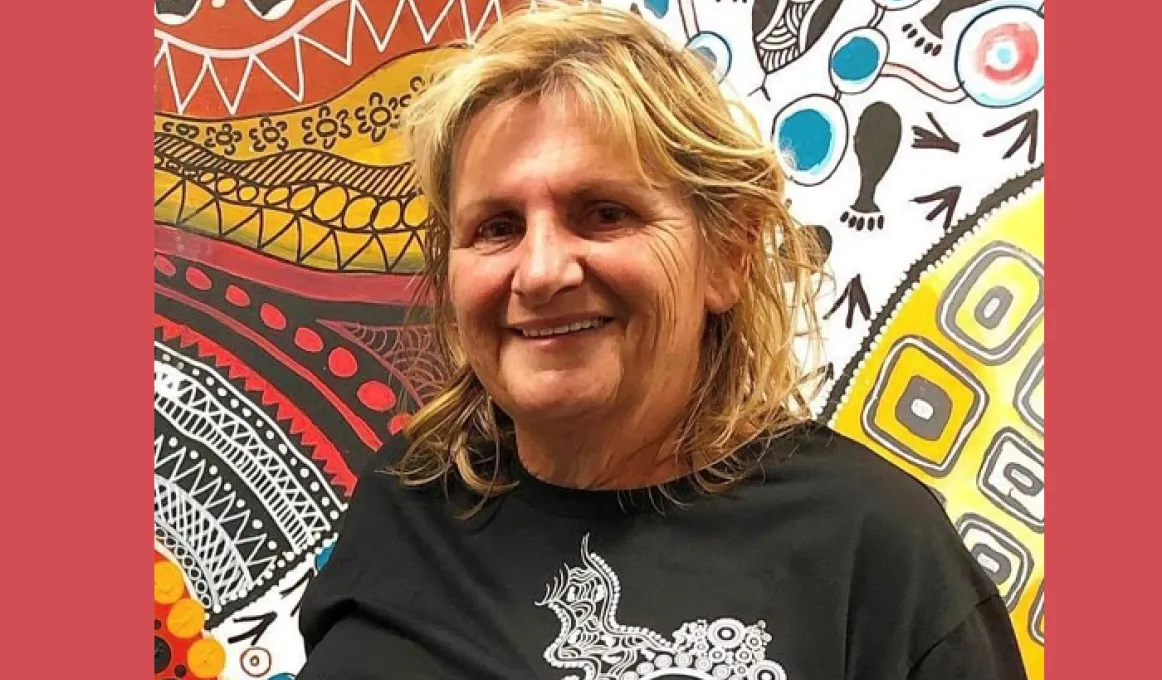 A mature woman with blonde hair wearing a dark shirt stands in front of a painting featuring traditional designs in multiple colours and features.