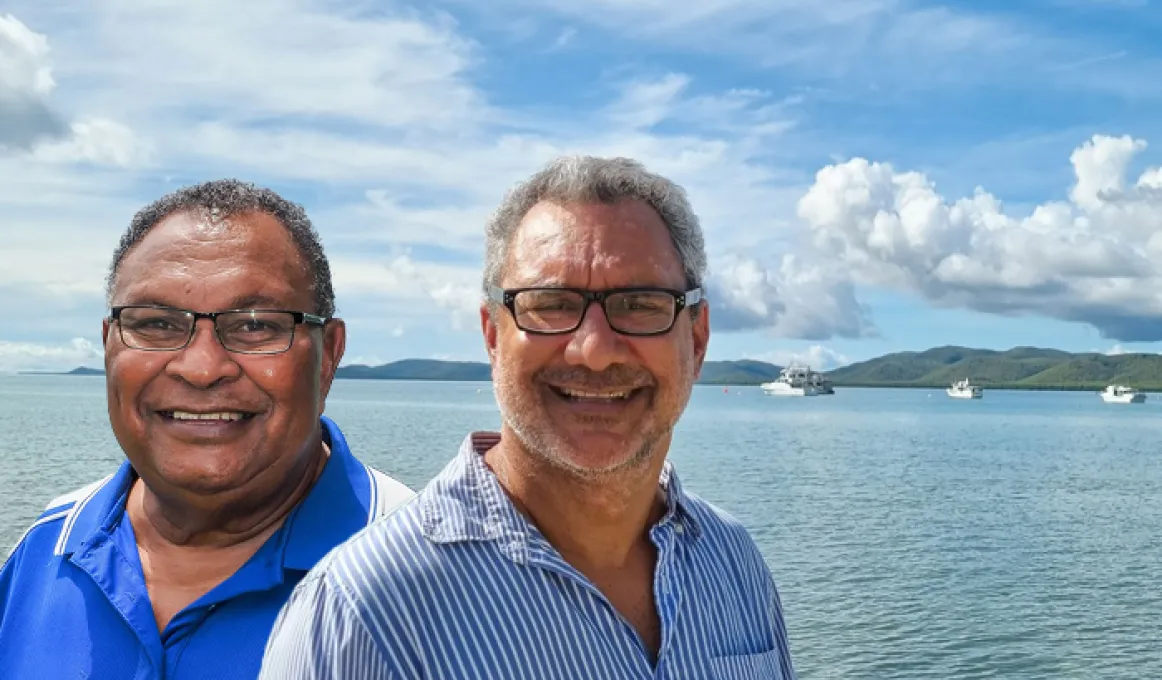 Head and shoulder photo of two men, both in blue shirts and wearing glasses. In the background is a large expanse of water, boats and hills.