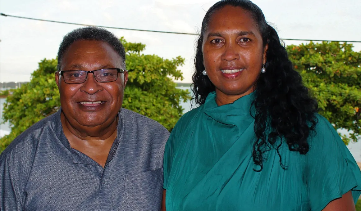 A man in a grey shirt and a woman in a green dress face the camera. In the background are trees and sky.