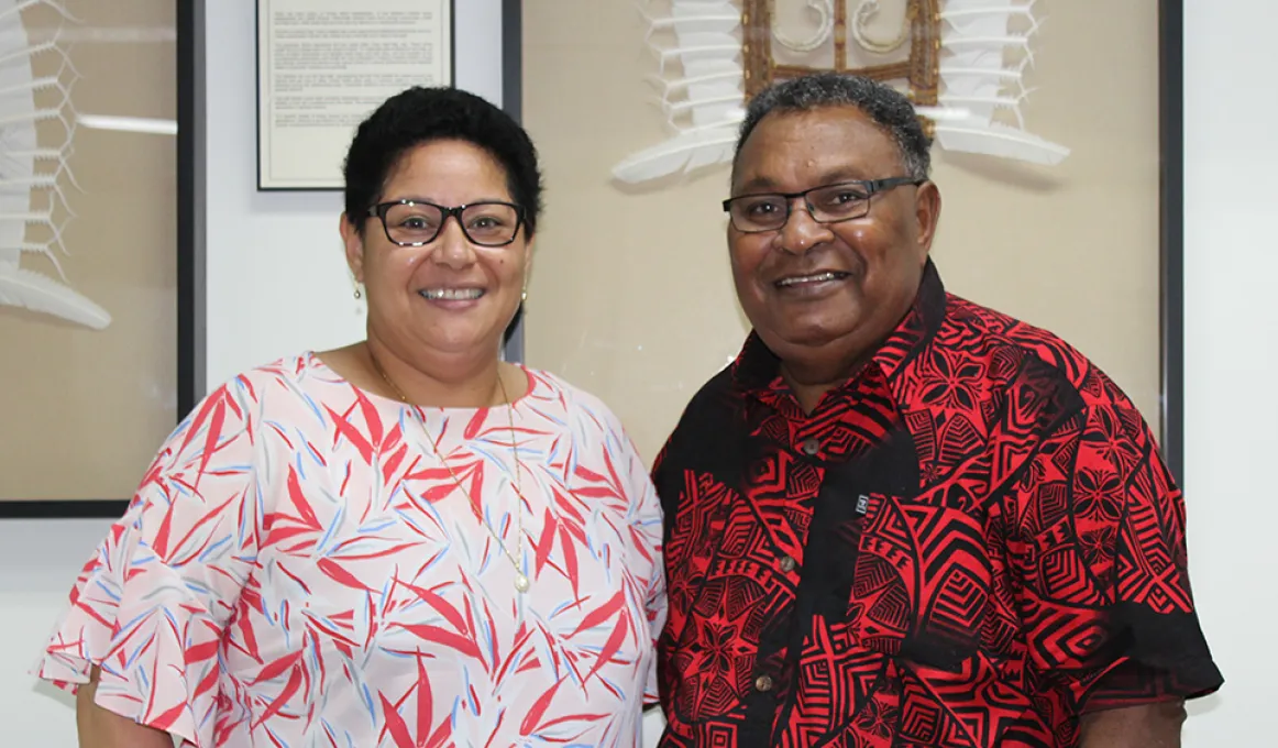 A woman in a pink floral shirt is standing next to a man in a red and black patterned shirt. Both the woman and man have short dark hair and black glasses.