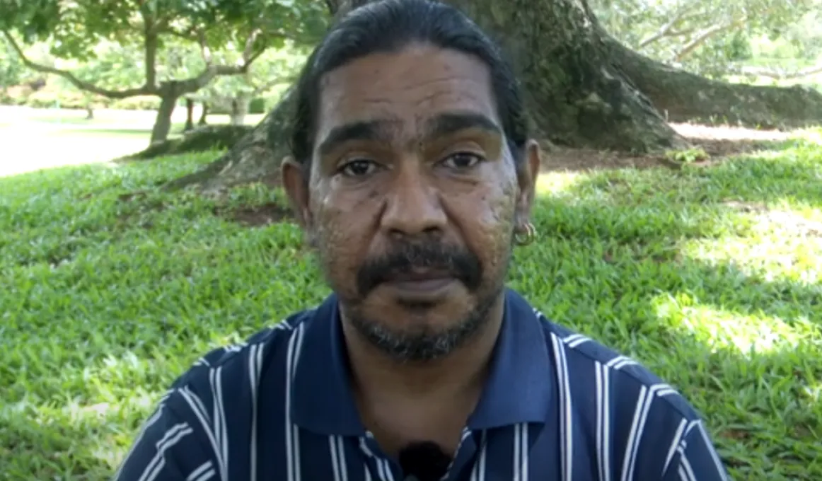 Man in dark blue and white striped shirt sits in grassed area in front of large tree.