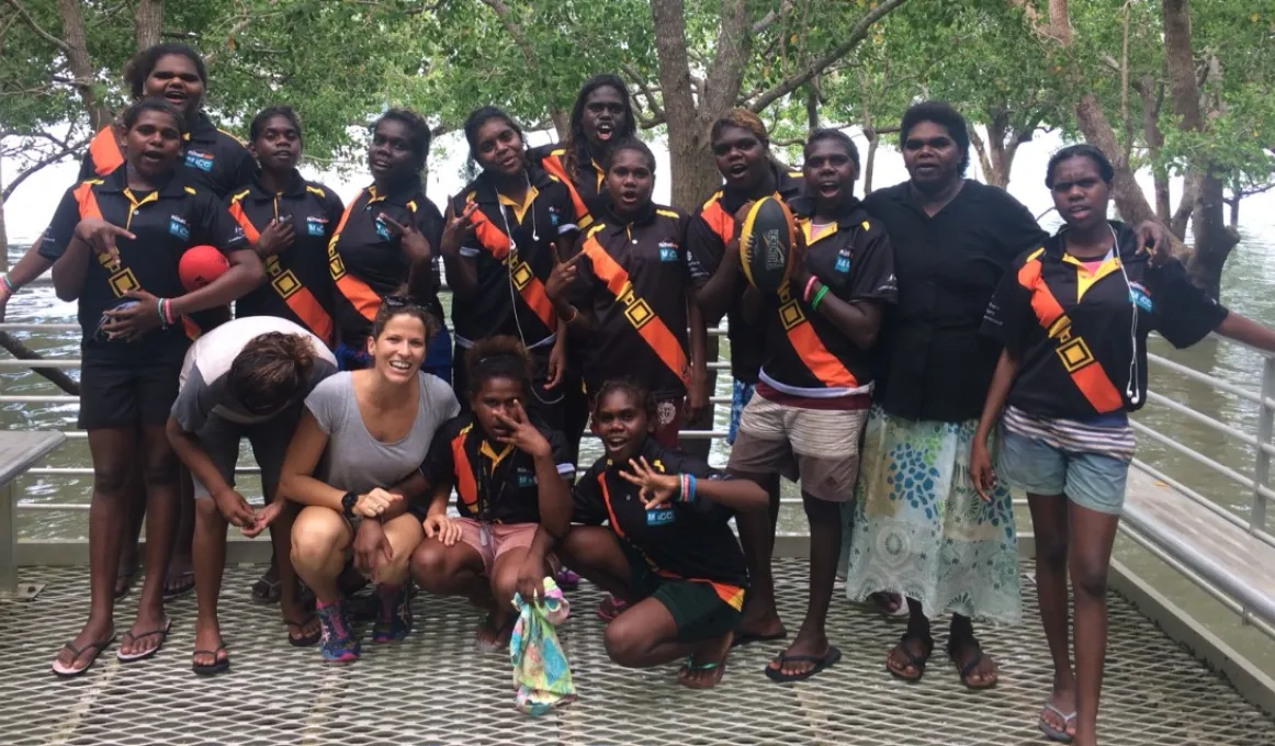 A group of young Indigenous women and their female supervisor standing near a body of water and trees.