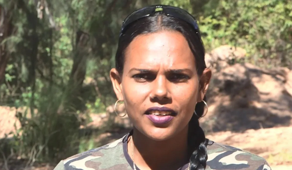 Aboriginal woman in cargo shirt and hair in plat looks at camera with trees in the background.