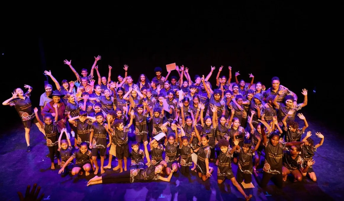 A large group of youth and a few adults stand or sit on a purple-lit stage. The background is black.