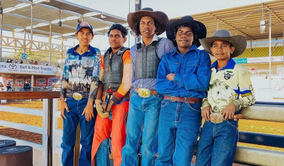 Five boys stand side by side leaning against a wooden fence. Behind them is a rodeo ring. Each of the boys is wearing jeans and a belt or work pants and collared work shirts. The boy on the left is wearing a cap and four of the boys aren't.