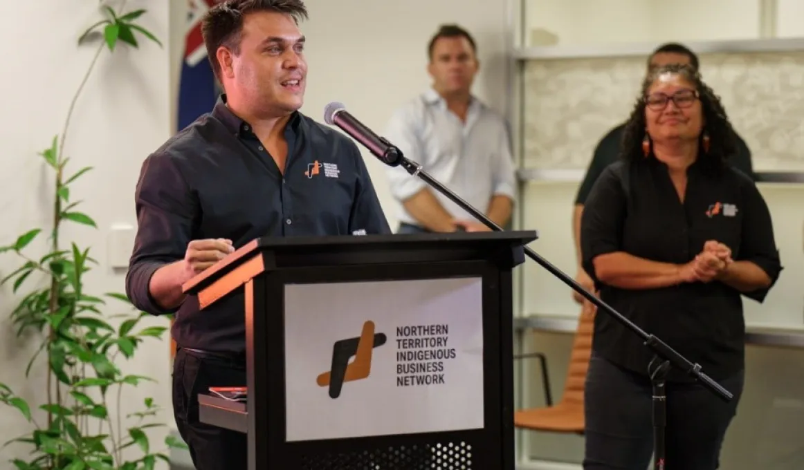 One person, dressed in black shirt with NTIBN logo is the focus of this image.  They are speaking into a microphone behind a lectern which displays a sign showing a logo which reads “Northern Territory Indigenous Business Network”.