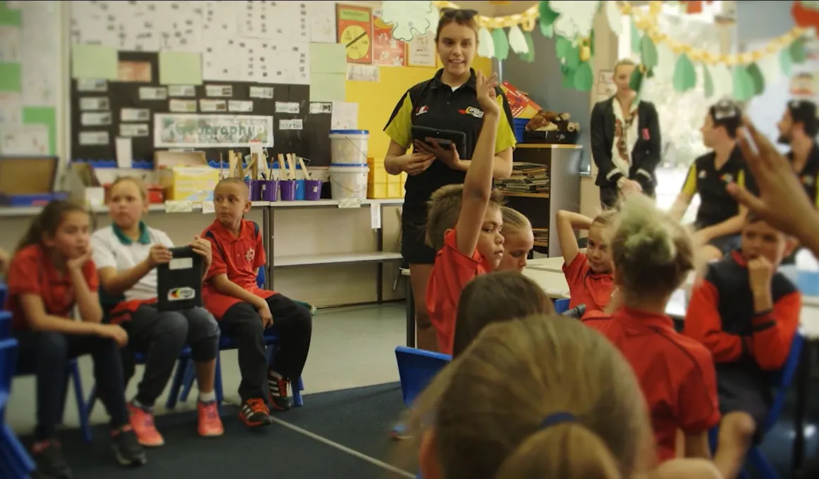 A young woman stands in a classroom teaching several Indigenous children seated on chairs. In the background are other young adults.