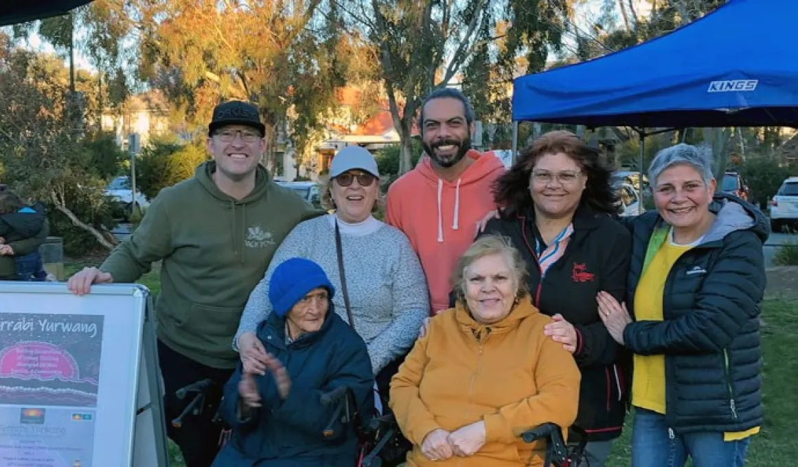 7 adults stand in a close group outdoors in front of a blue marquee tent. Each of them has their hand on the shoulder or arm of each other. The man on the left has his hand on a whiteboard sign that has a poster for Yerrabi Yurwang.