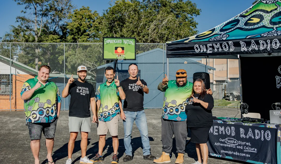 Six people in casual wear stand shoulder to shoulder on a hard surface outdoors. Next to them is a table with a banner displaying the words OneMob Radio. In the background is a building.