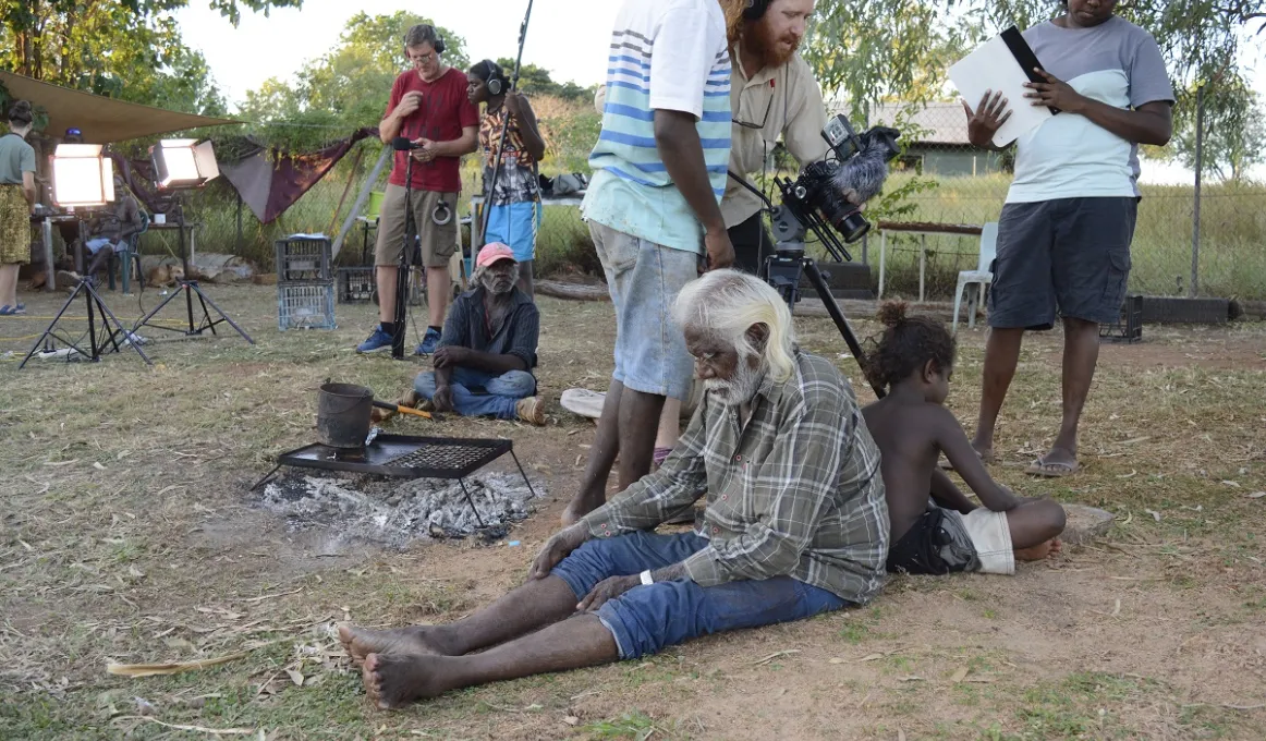 Elderly Aboriginal man sits back to back with young Aboriginal boy on grassy ground. Immediately behind them are three film-makers and further back are more Aboriginal and non-Aboriginal film makers another Aboriginal actor, lights, trees and grass.