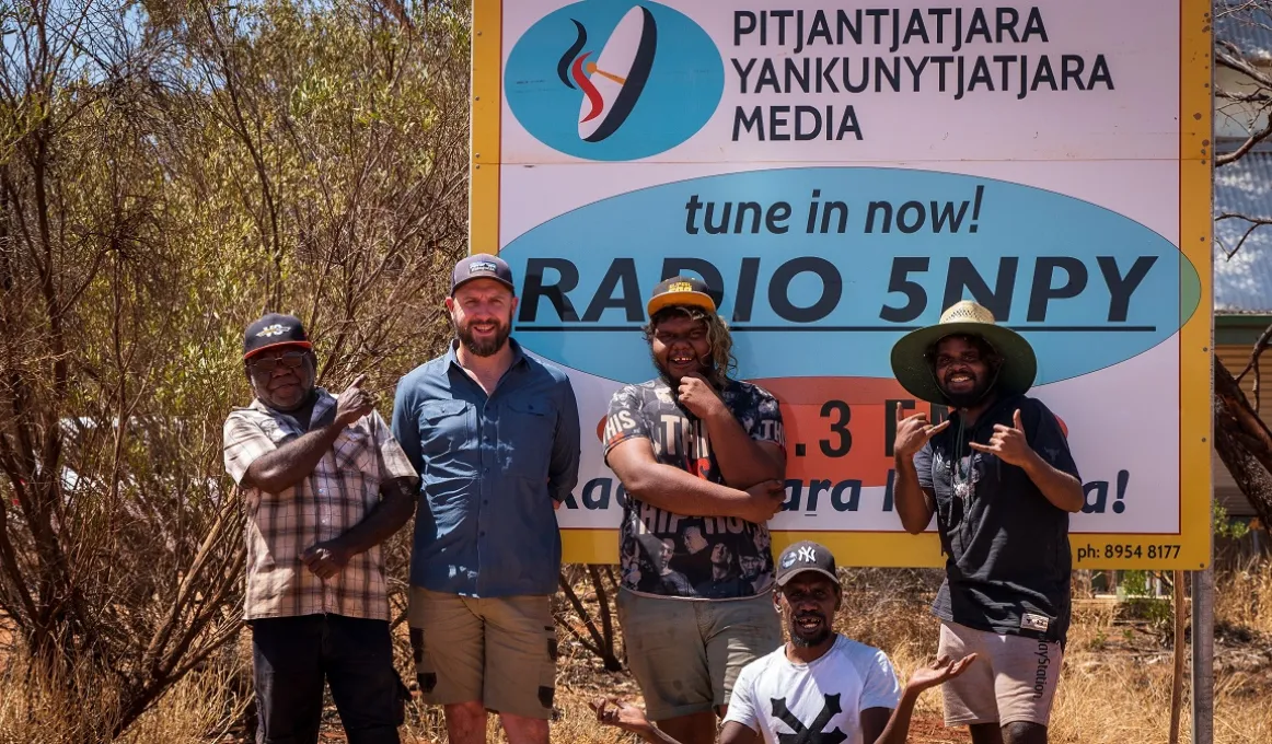 Five men in casual wear stand or sit in front of a large multi-coloured sign with a logo at top left and the following text: Pitjantjatjara Yankunytjatjara Media. Tune in now Radio 5NPY. In the background are trees, grass and a building.