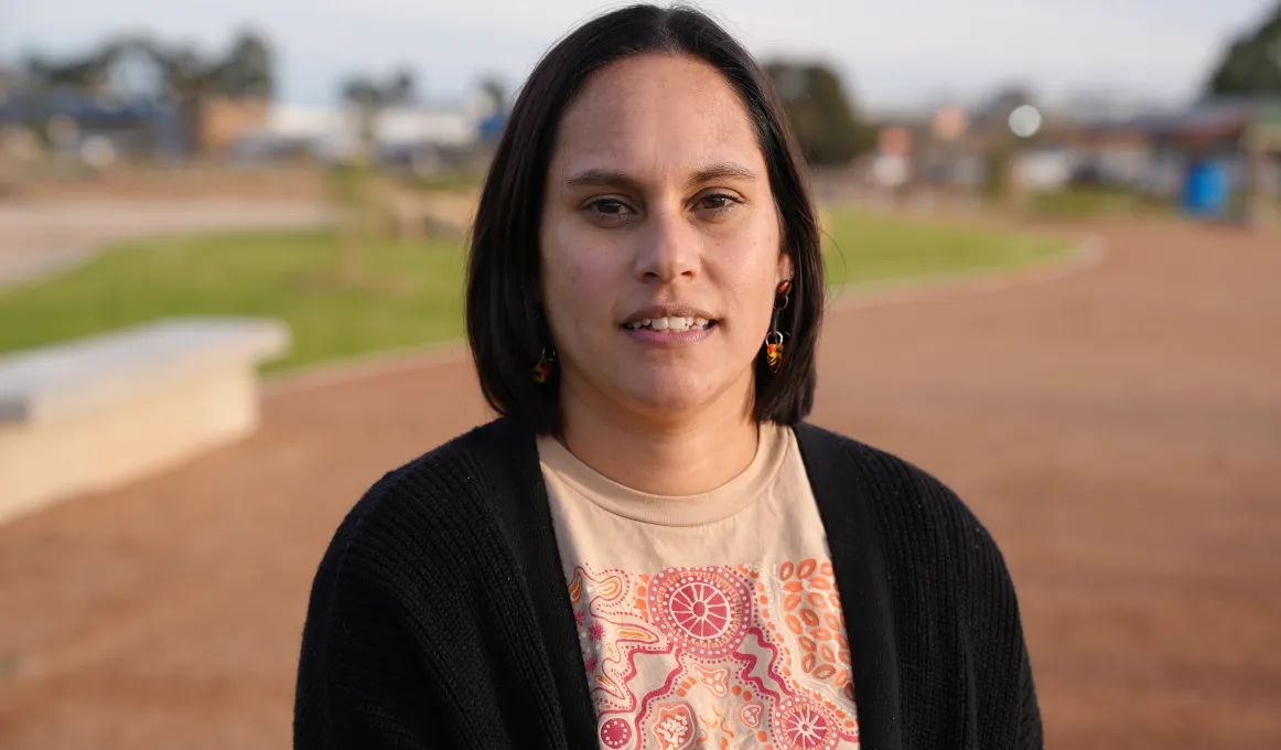 A woman with dark shoulder length hair and wearing a black top over a colourful top with traditional designs faces camera. In the background is brown soil, green grass and some buildings.