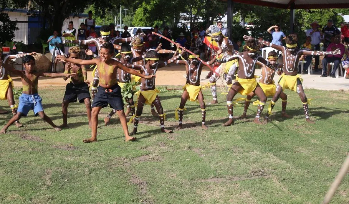 A group of Indigenous children with white ceremonial paint on their bodies wearing shorts or yellow coverings dance on green grass watched by a crowd of onlookers.