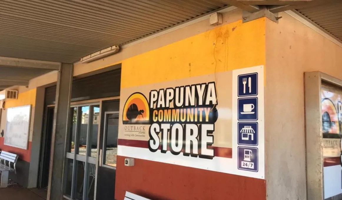 We see the corner of a shop and the eaves at the top. At the shopfront are two silver coloured benches. There are various signs on the walls including the words: Panunya Community Store.