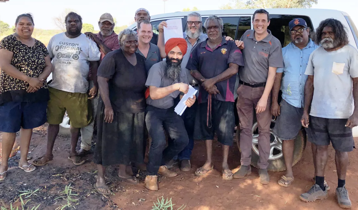 There are 12 adults standing together and facing the camera. They are outside on a sunny day, standing in the shade, in front of a white van.