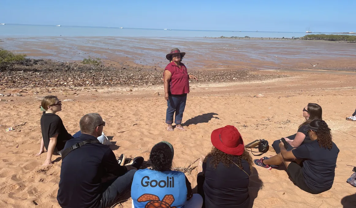 A group of people sitting on a beach listening to a woman wearing a red shirt and a broad brimmed hat.