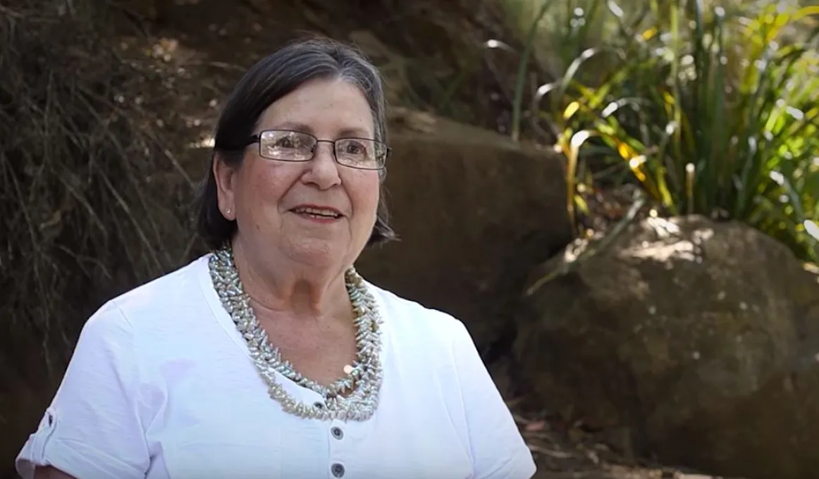 Elderly woman with dark hair wearing glasses, multiple necklaces and white dress stands in front of rocks and green foliage.