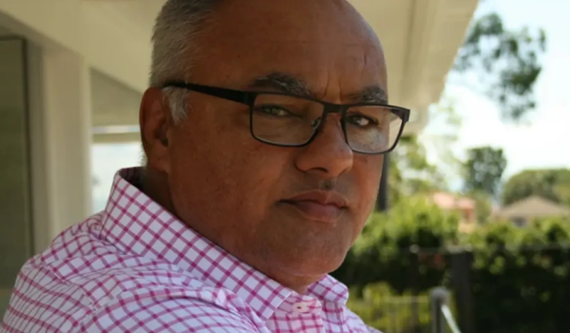 A man wearing a red checked shirt and black rimmed glasses looks at camera. In the background are bushes, a fence and some grass. Above him is a ceiling.