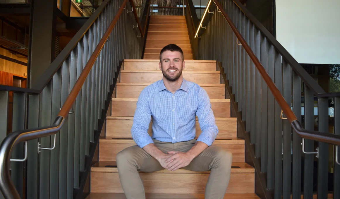 Young adult man wearing blue shirt and light brown pants sits on wooden staircase with grey railings to balustrade.