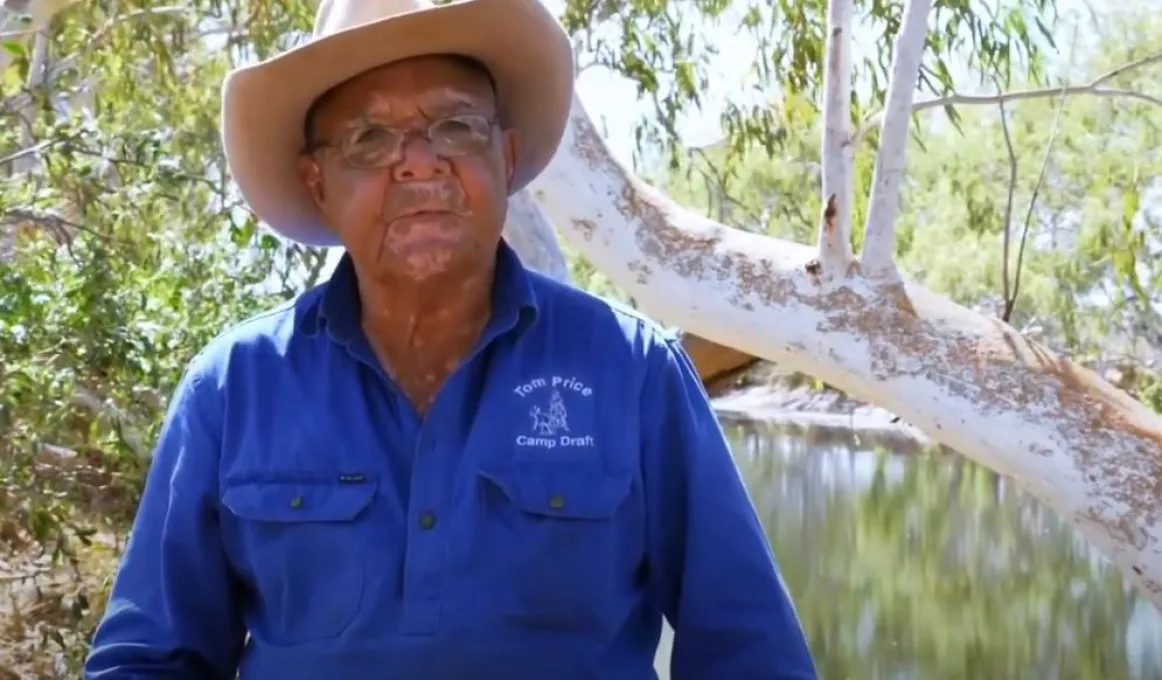 Mature aged man in blue shirt and beige brimmed hat stands in front of the bough of a tree. Behind him are more trees and a river.