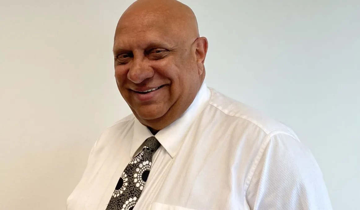 Elderly and bald man in shirt and tie is in front of a pale background.