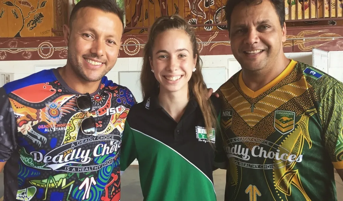 Two men and a young woman who stands between them face the camera. They all wear colourful shirts and in the background is a wall painted with Indigenous designs.