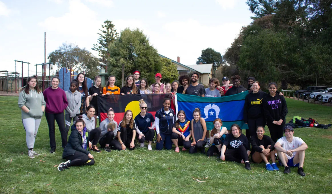 Group of Indigenous youth sit or stand on grass, some holding the Aboriginal flag and Torres Strait Islander flag. In the background is a building, cars and trees.