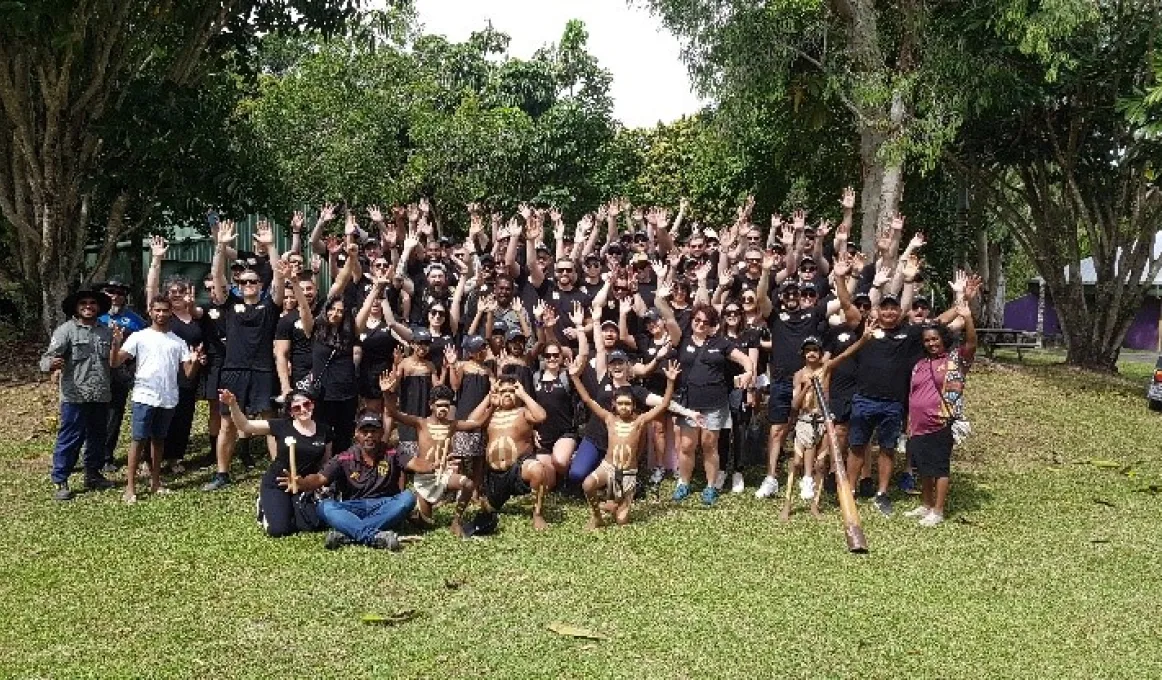 A huddled group of staff in Yarrabah, covered in Indigenous paint, standing with their hands in the air. Some staff are holding Indigenous instruments.