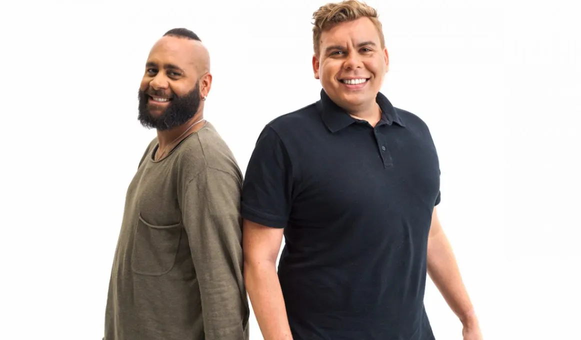 Two men are standing back to back smiling at the camera. The man on the left is wearing a brown long sleeved t-shirt and has a beard and mohawk. The man on the right has a dark polo and light, short wavy hair.