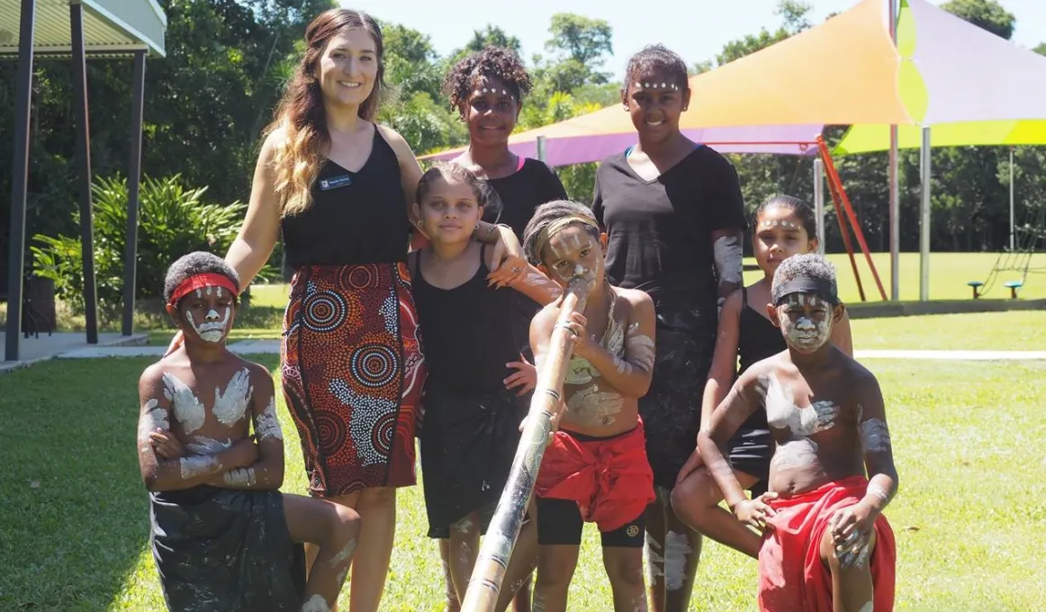 An adult woman and seven Indigenous youth stand or kneel on a lawn. To the left is a building and in the background a canopy. Some of the youth wear paint in traditional patterns and wear traditional clothing while one holds a didgeridoo.