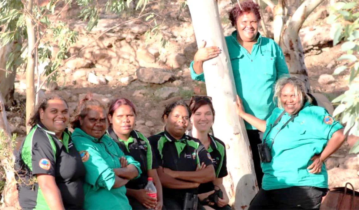 Seven Aboriginal women in ranger uniforms stand next to a white bark tree. In the background is a rocky slope and more trees.