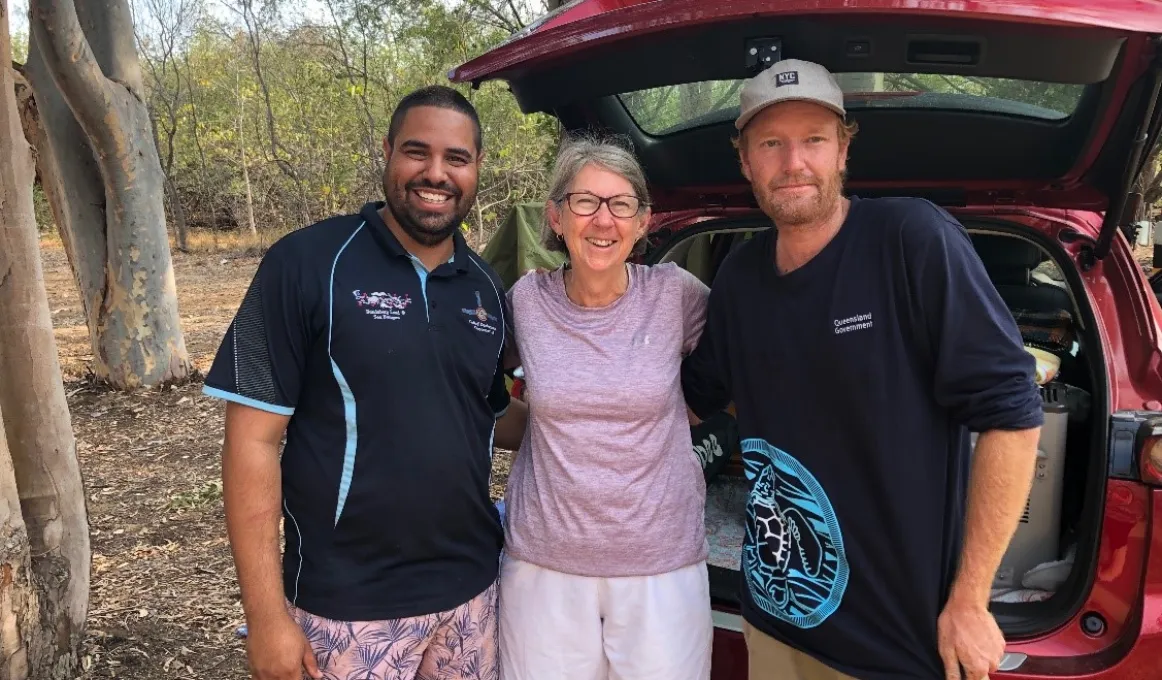 A mature woman in lilac coloured top and white shorts stands between, and arm in arm, with two men dressed in blue shirts. Behind them is a red car with its rear door open. In the background are trees and bare soil.