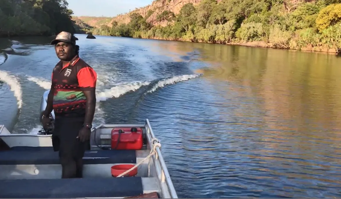 A young man stands up and steers a boat on a river. In the background is water, vegetation and a cliff face.