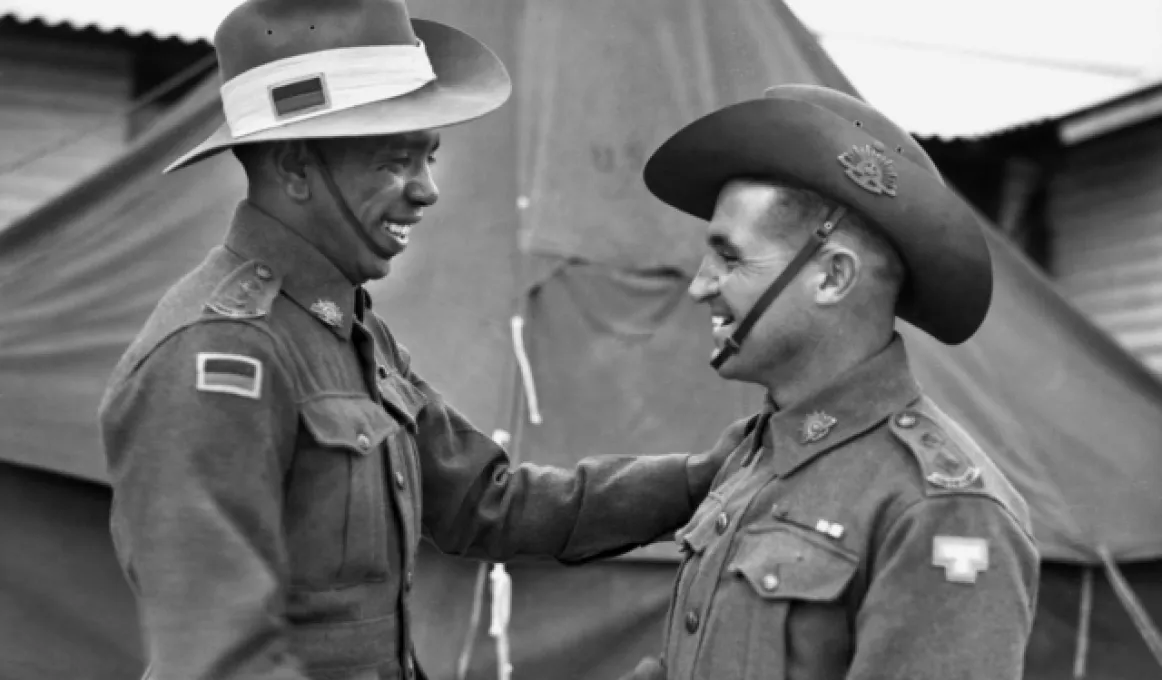 A smiling man in army uniform shakes hands with another smiling man also in uniform. In the background is a large tent and a building.