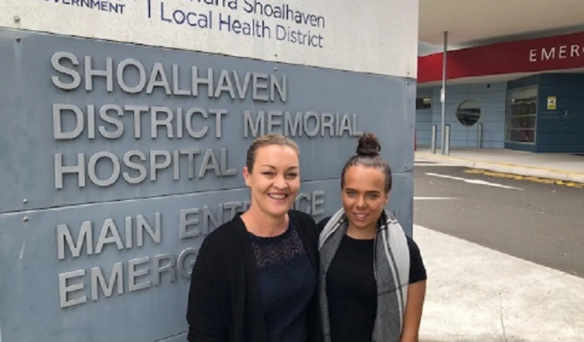 Two women stand in front of a large sign featuring the words Shoalhaven District Memorial Hospital. The woman at left wears a dark cardigan and dress and the one on the right wears a dark dress with a grey and white scarf about her neck.