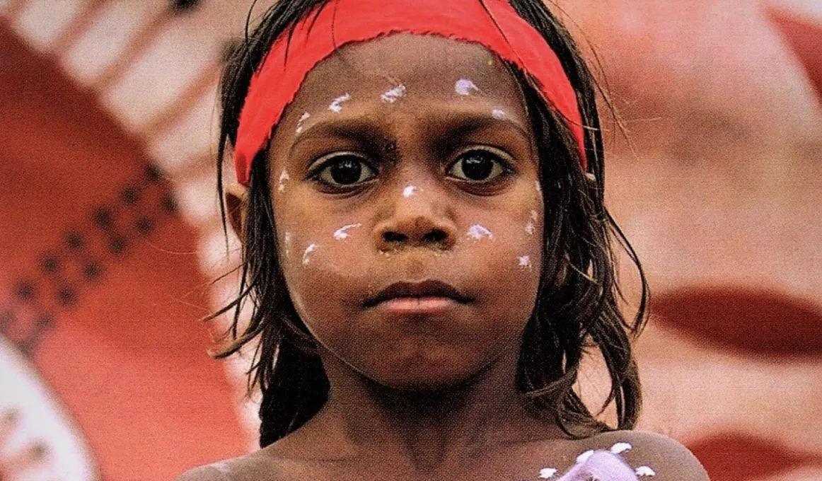 Aboriginal boy in body and face paint standing in front of Aboriginal artwork