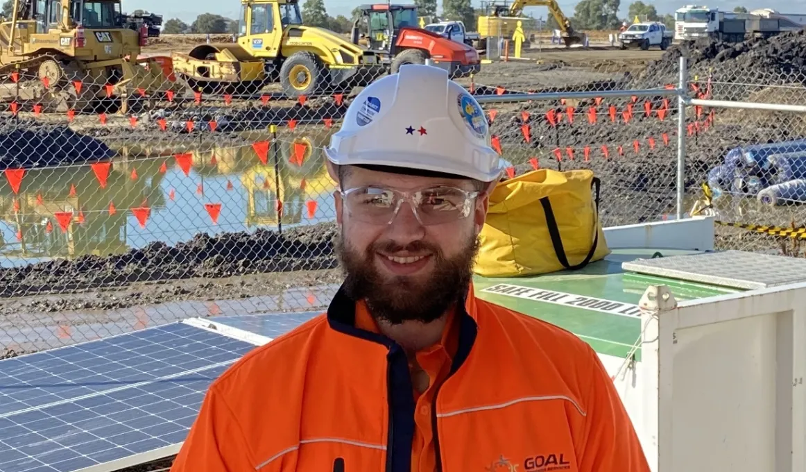 A young adult male in orange work-wear and white helmet stands in front of a white container and solar panel. In the background are large earthmoving vehicles and trucks.