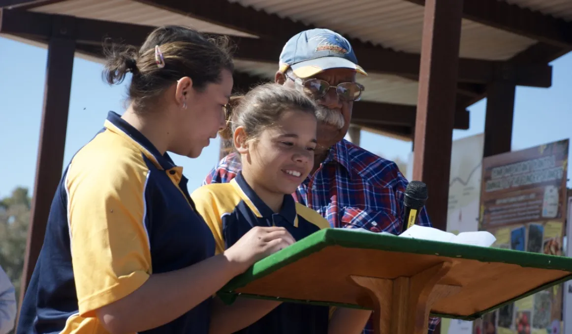 Two young Aboriginal girls dressed in blue and yellow polo shirts stand at a podium with an elderly Aboriginal man in blue cap and blue and red shirt. In the background is a small metal shelter.